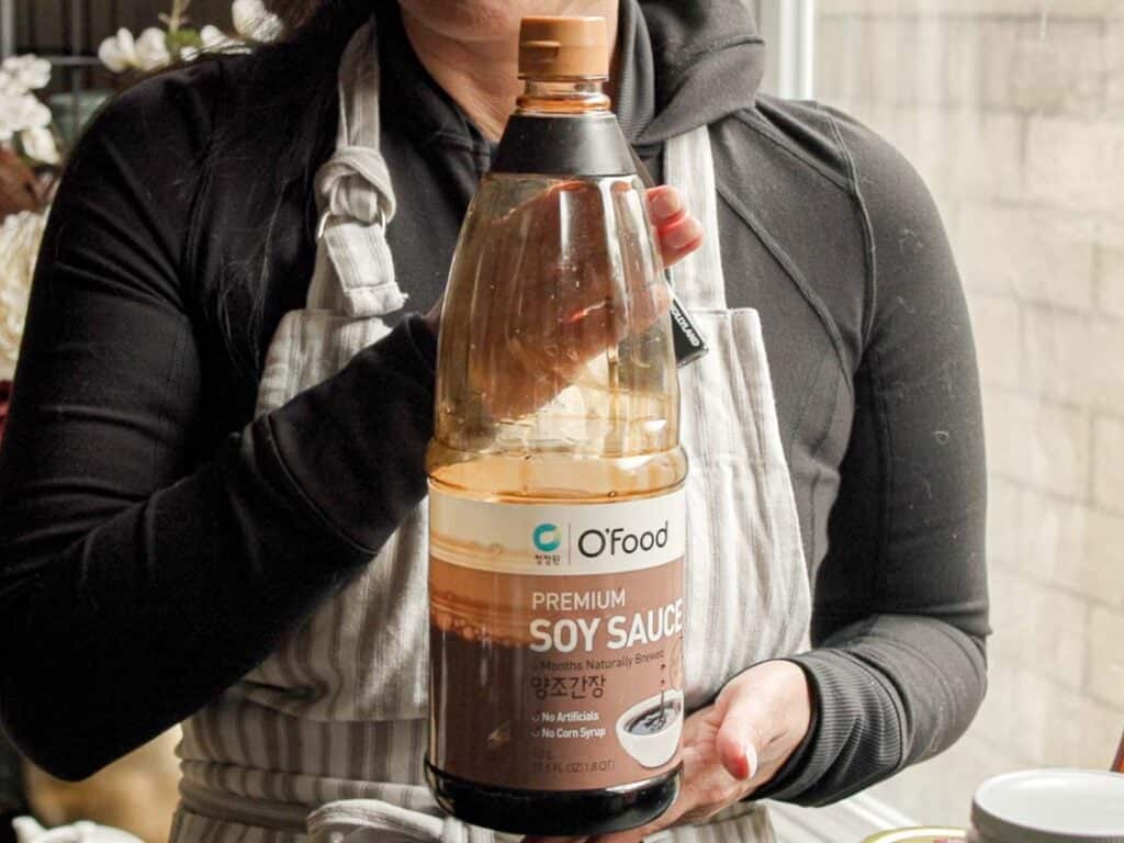 A person in a gray apron is holding a large bottle of OFood Premium Soy Sauce. The label includes text in Korean and English. The person is standing near a window with a neutral background.